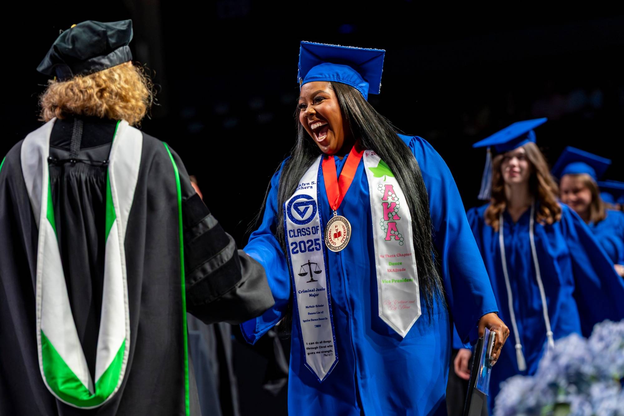 A grad smiles while receiving her diploma cover and shaking hands with a faculty member